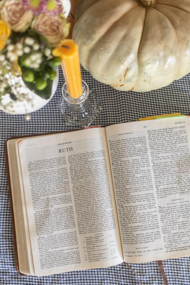 bible open to ruth 1 on blue and white gingham tablecloth with a rose floral arrangment in a white pitcher, a yellow candlestick and a green pumpkin centerpiece framing the top of the photo