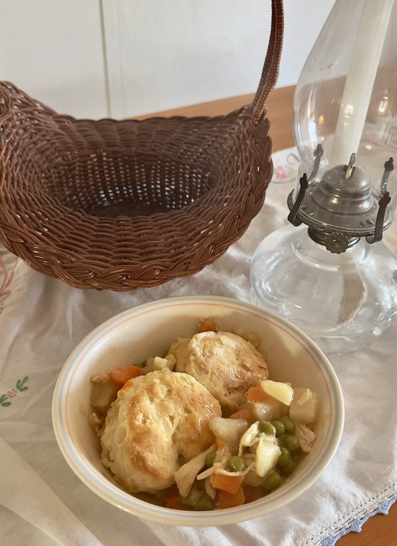 bowl of gluten free chicken pot pie with a gluten free biscuit topping on vintage tablecloth with a basket and antique oil lamp