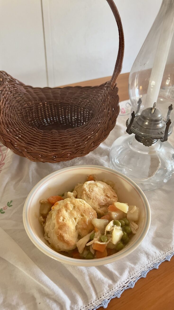 bowl of gluten free chicken pot pie with a gluten free biscuit topping on vintage tablecloth with a basket and antique oil lamp