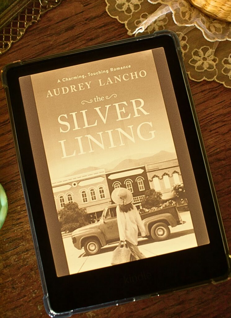 Kindle E-Reader with the book the silver lining by audrey lancho pulled up, laid on a wooden dresser top with a lace doiley and vintage metal picture frame in the background