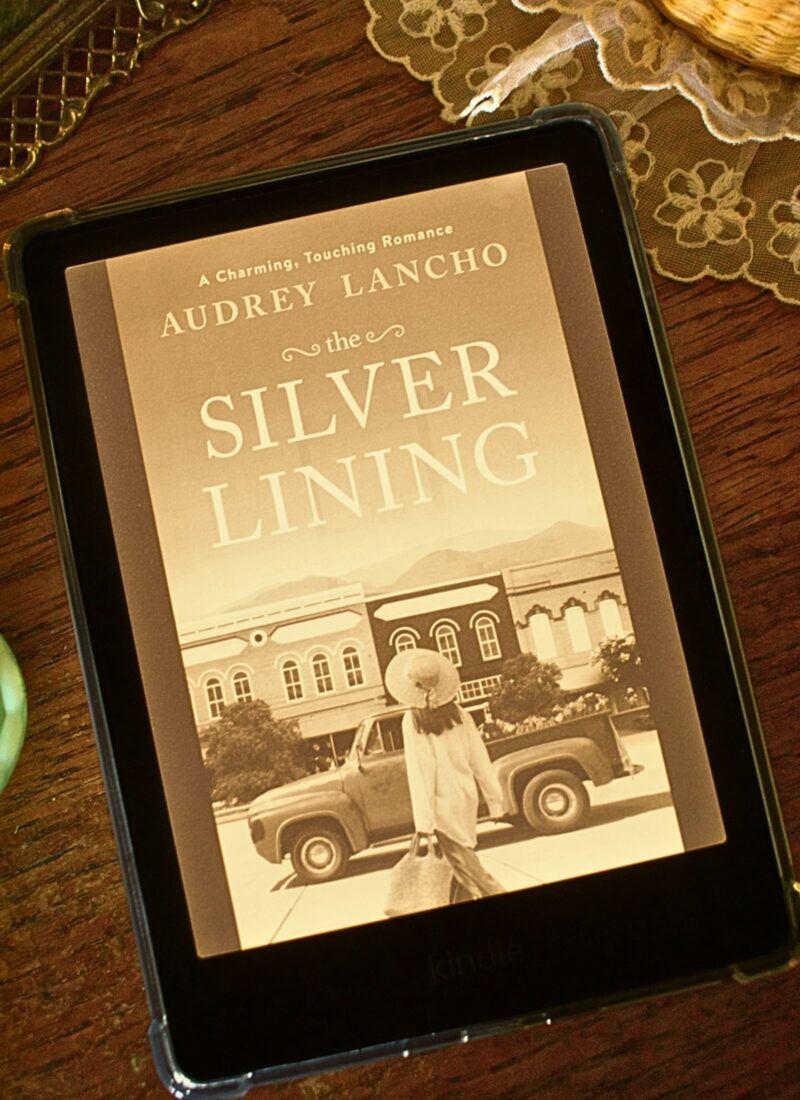 Kindle E-Reader with the book the silver lining by audrey lancho pulled up, laid on a wooden dresser top with a lace doiley and vintage metal picture frame in the background