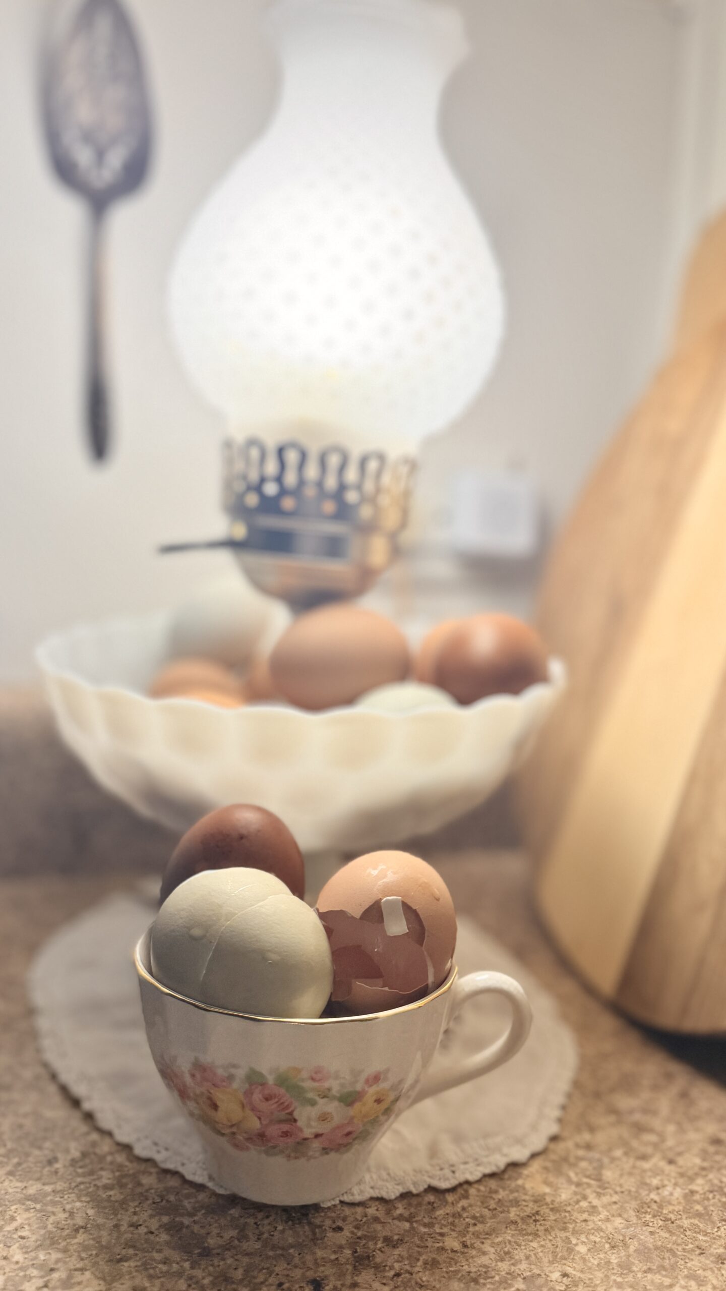 Green and brown egg shells stacked in an antique teacup in front of an antique milk glass lamp full of eggs.