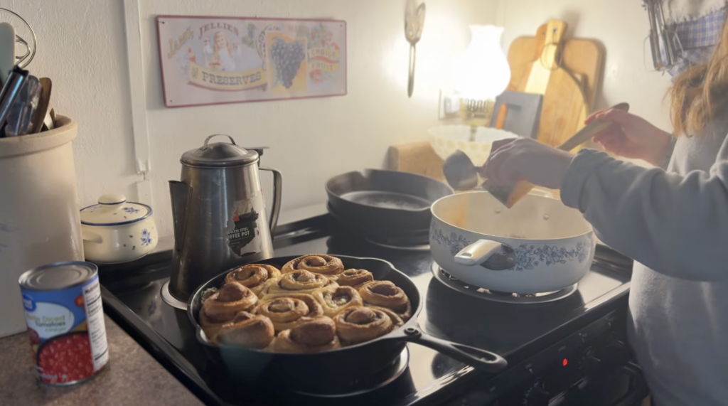 pioneer woman blue and white pot on stovetop while adding chili seasoning with cinnamon rolls sitting beside in a cast iron pan
