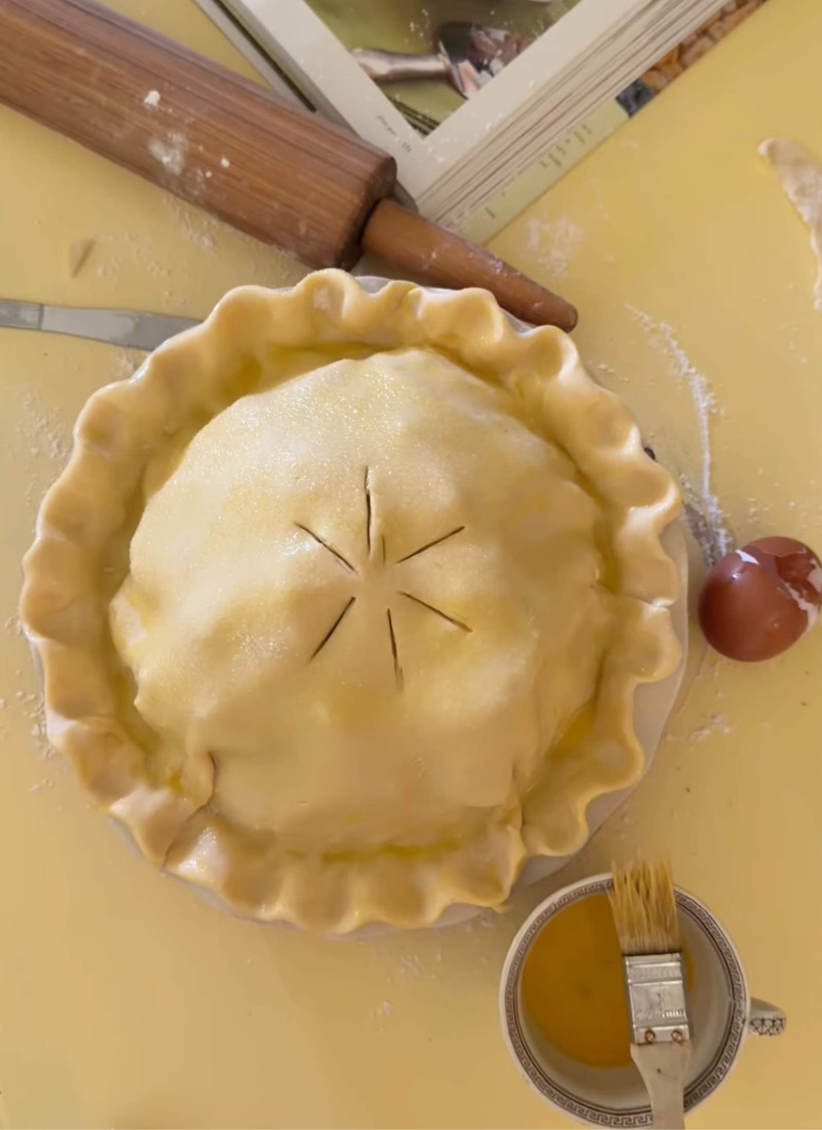 deep dish pie crust with crimped edge ready for the oven sitting on a butter yellow table with an old wooden rolling pin, southern desserts cookbook and a vintage teacup with leftover egg wash sitting around it.