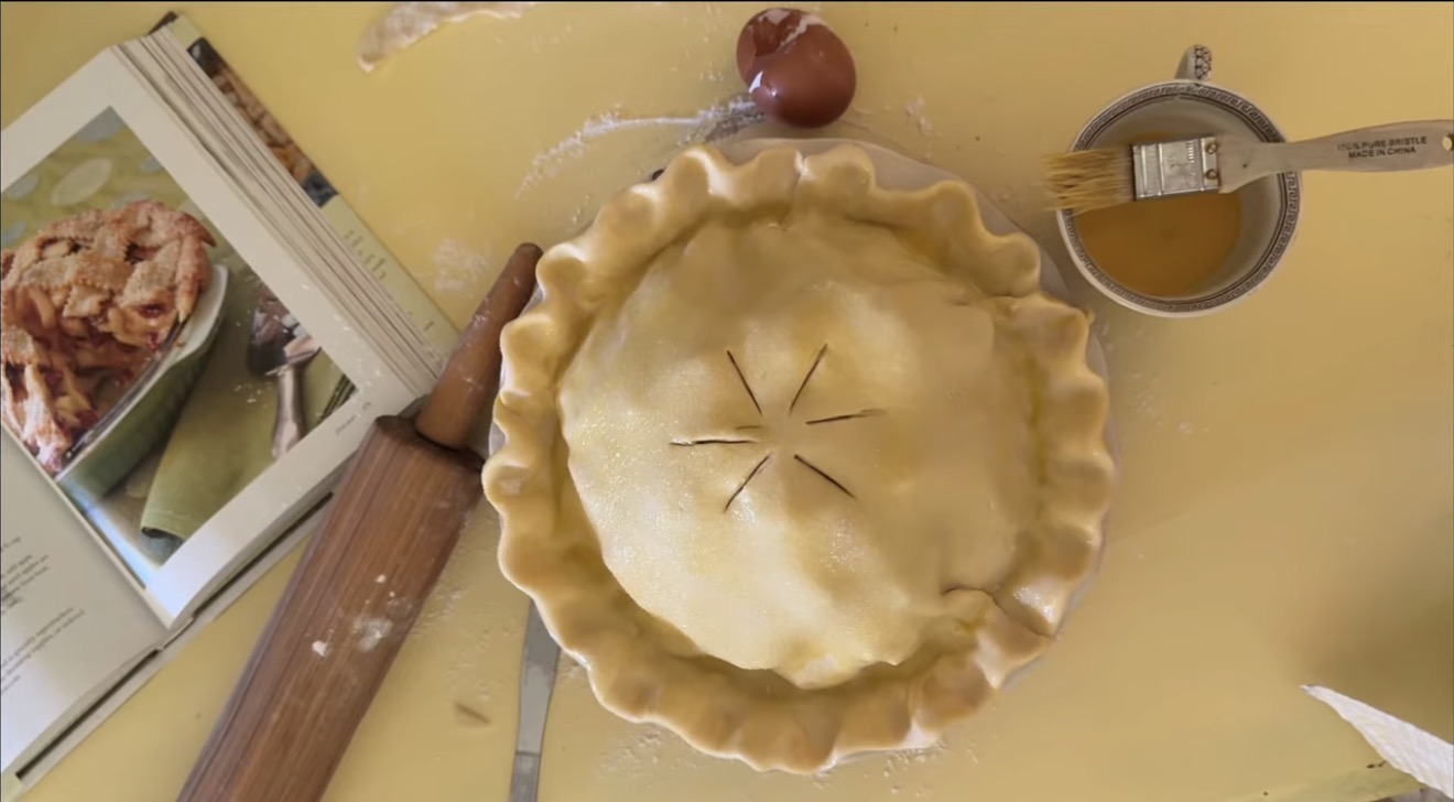deep dish pie crust with crimped edge ready for the oven sitting on a butter yellow table with an old wooden rolling pin, southern desserts cookbook and a vintage teacup with leftover egg wash sitting around it.